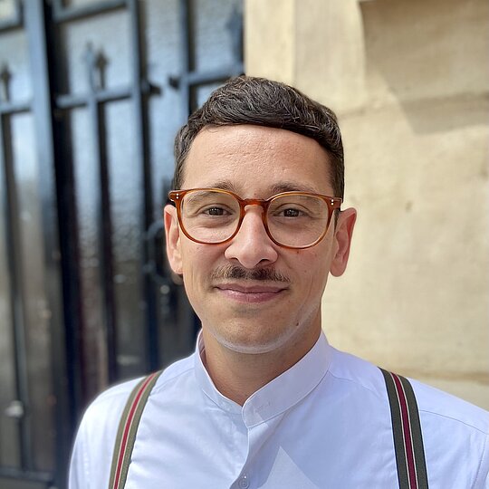 A man with a mustache and brown glasses wearing a white shirt.