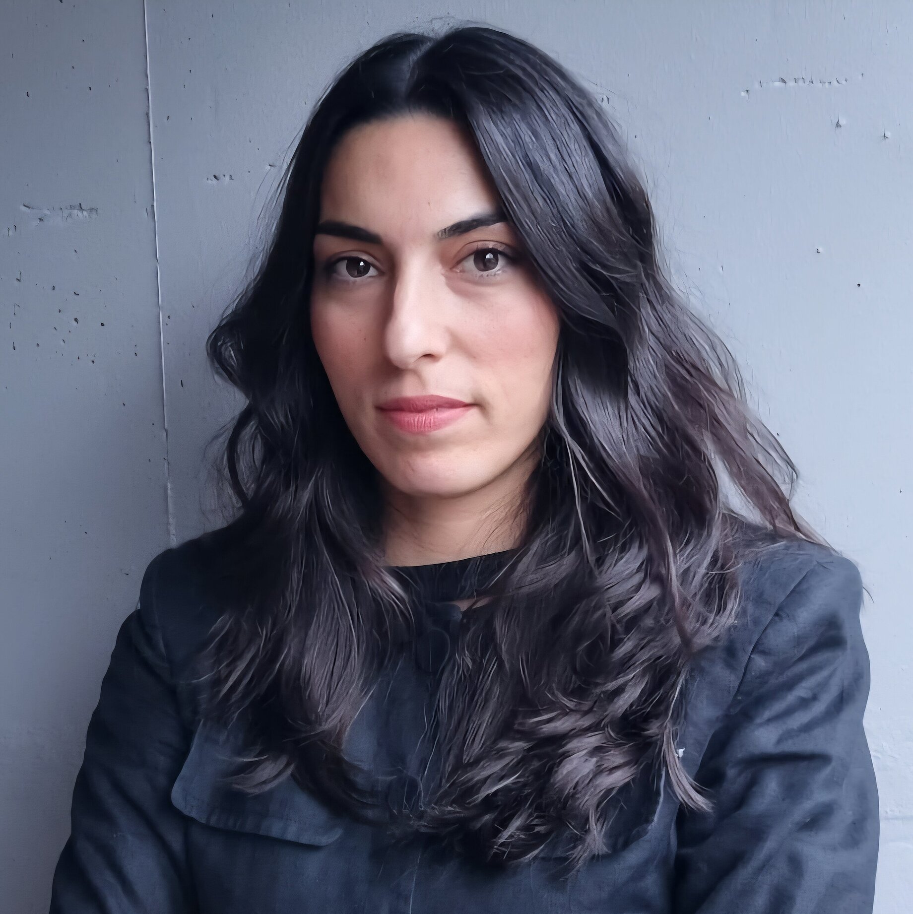 A woman with wavy black hair wearing a black jacket. She stands in front of an industrial wall as background
