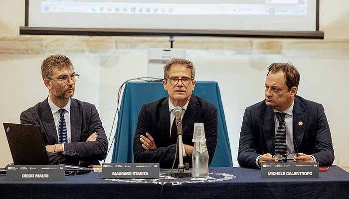 Three men are sitting on a panel, the man in the middle is speaking