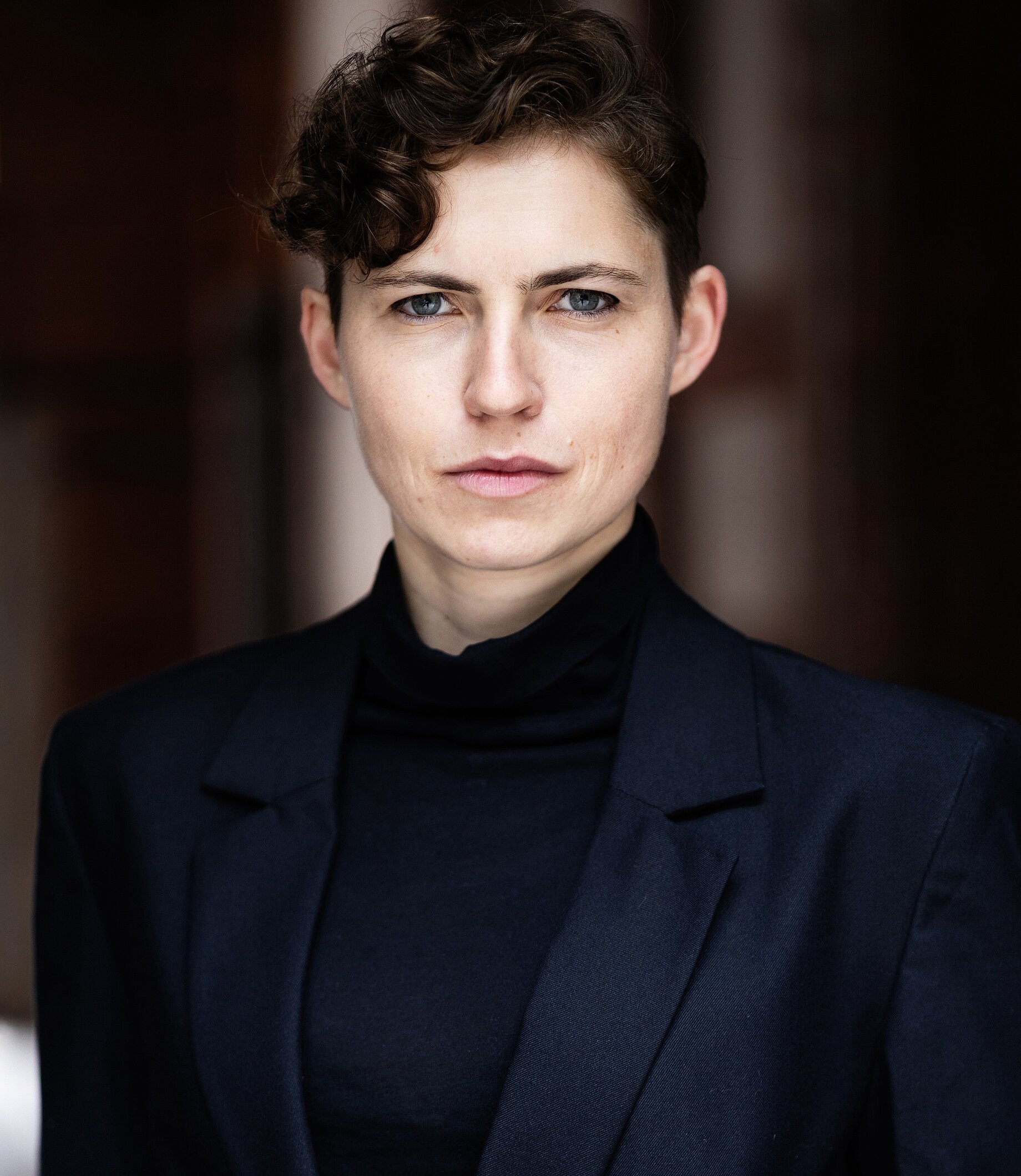 A person with short hair wearing a navy blue jacket on top of a black top.