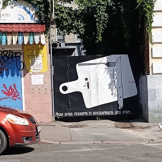 Street view of a mural, on it is a cutting board, with a knife and fork perpendicular to one another, with the captioned text below.