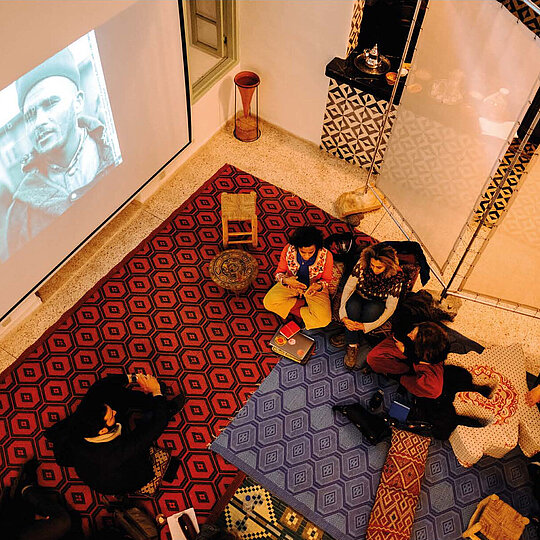 Group of people sitting on patterned cushions and rugs in a room watching a black-and-white video projected on a wall.