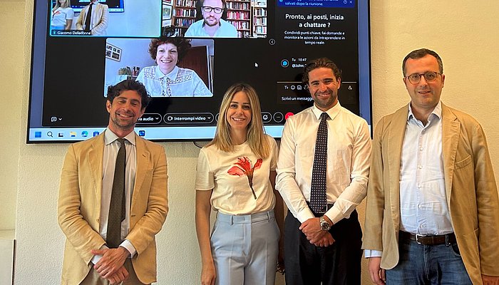 Photo of four panelists standing in front of a big screen with further people in a Zoom call