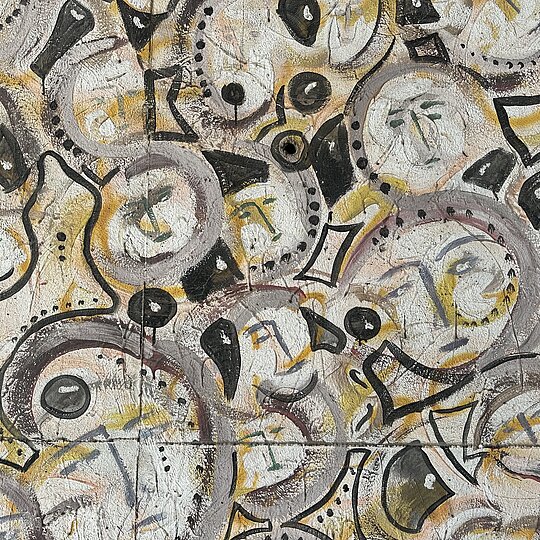 An abstract mural with multiple circular faces and geometric shapes in black, gray, and yellow on a textured wall.
