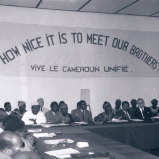 Old black and white photograph of a group of people sitting on a conference table under a sign saying "How noice it is to meet our brothers! Vive le cameroun unifié."