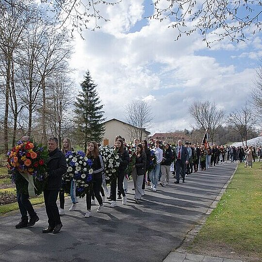 People walking in a line carrying wreaths with flowers on the grounds of the former concentration camp.