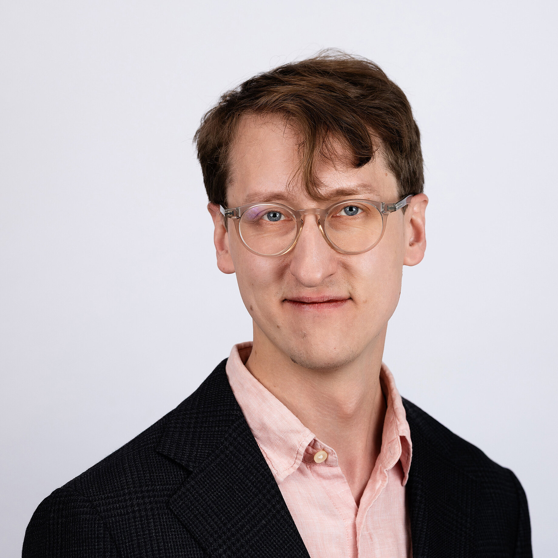 A man with short light brown hair wearing glasses and a black jacket on a pink shirt