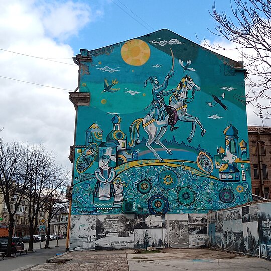 A bright blue mural on the side of a large building, In the centre of the mural, a man is depicted sitting upon a horse with a sword raised towards the sky.